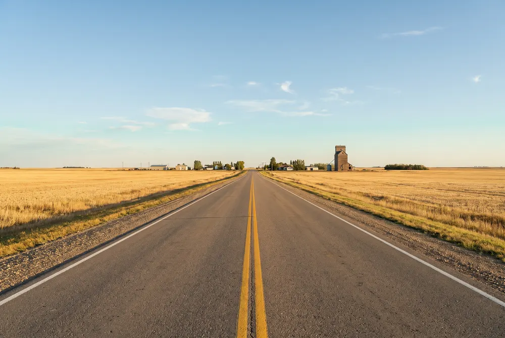Generated illustration: a straight two-lane prairie highway through golden fields toward a small town with a grain elevator